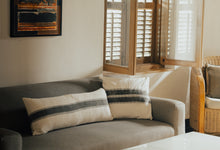 Load image into Gallery viewer, Charcoal stripe band printed in the middle of the extra long lumbar cotton cushion cover on an ivory base fabric. Sitting on a couch next to a charcoal printed cotton cushion cover