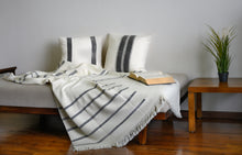Load image into Gallery viewer, Hand woven woollen throw blanket in a charcoal grey and ivory colour placed next to two block printed charcoal cushions on a day bed
